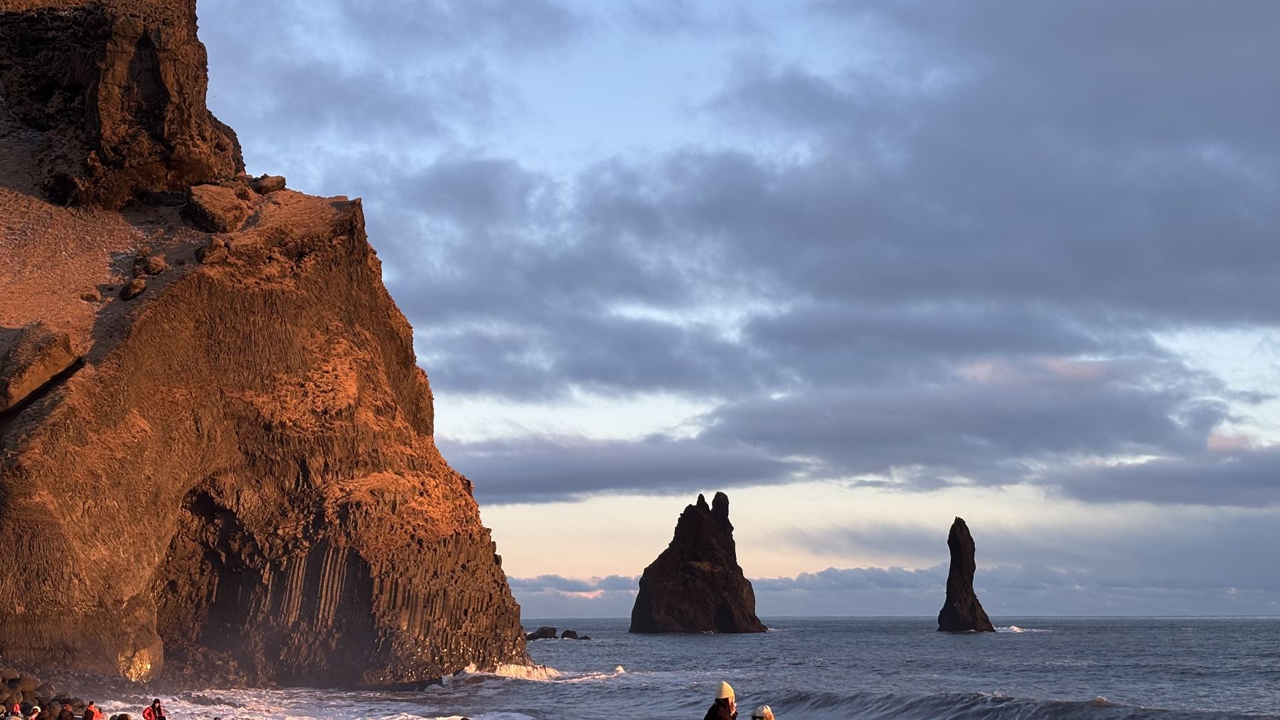 Acantilado de basalto iluminado por el sol bajo y los farallones de Reynisdrangar en el mar